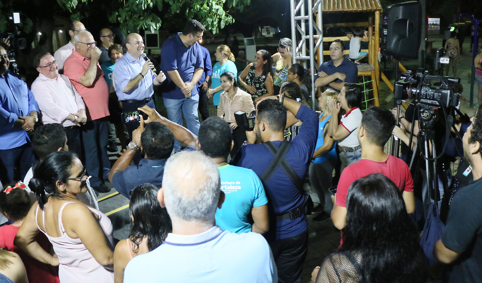 prefeito falando ao microfone em cima de palco com várias pessoas ao redor e em frente assistindo